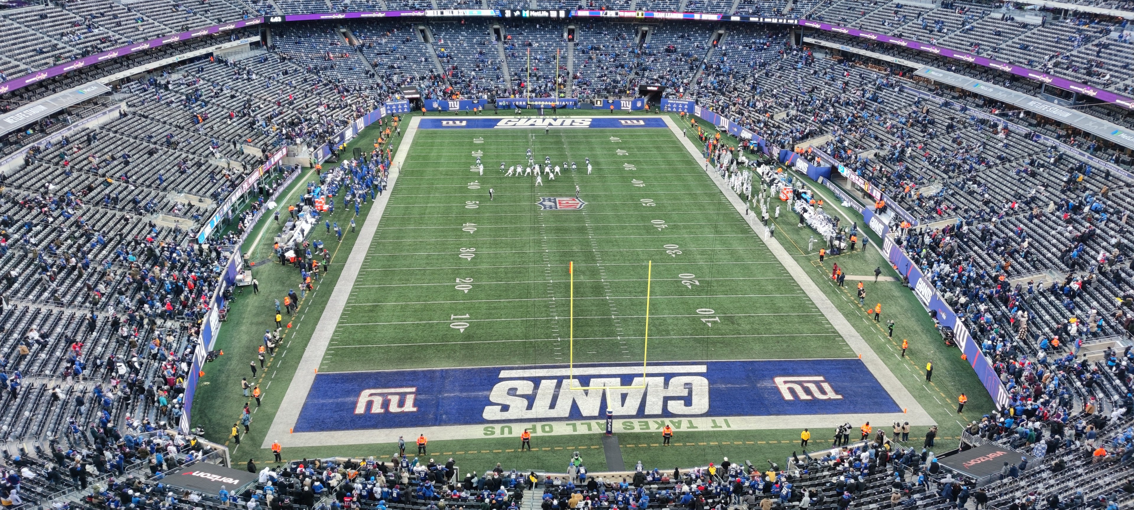 Ein volles Fußballstadion mit Spielern auf dem Feld, Torpfosten und Textanzeigen, aus einer erhöhten Perspektive betrachtet.
