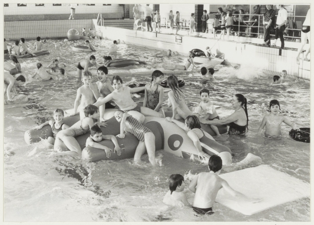 Schwarzes und weißes Bild von Kindern, die in einem Schwimmbad spielen, einige sitzen auf aufblasbaren Gegenständen und andere stehen auf dem Poolboden, mit einer Wand und einem Geländer im Hintergrund und Text unten.