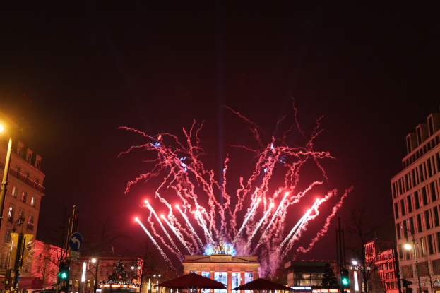 Eine belebte StraßenSzene in Berlin an Silvester, voller Menschen, Fahrzeuge und erleuchtet von Feuerwerk und Gebäudelichtern, die eine festliche Atmosphäre schaffen.