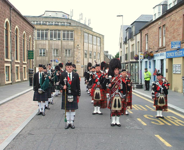 Eine Gruppe von Menschen, einige stehen auf der Straße und andere spielen Musikinstrumente, mit Gebäuden, Plakaten, Polen, Bäumen und Reifen im Hintergrund.