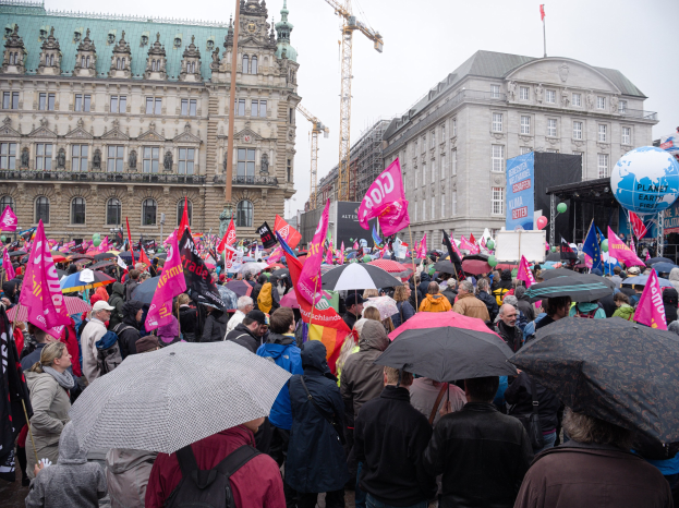 Eine große Gruppe von Menschen nimmt an einer Frauen-Demo in Berlin teil, mit Schirmen und Plakaten, Ballons und Stangen im Vordergrund und einem Gebäude im Hintergrund.