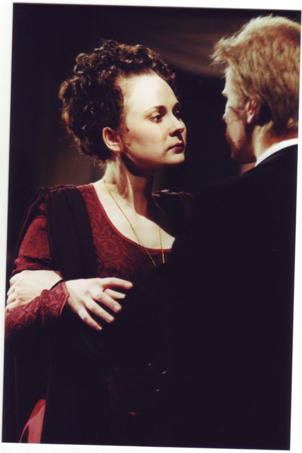 Eine Frau in einem roten Kleid mit einer Kette steht neben einem Mann in einem schwarzen Anzug, der sie liebevoll ansieht, vor einem dunklen, romantischen Hintergrund.