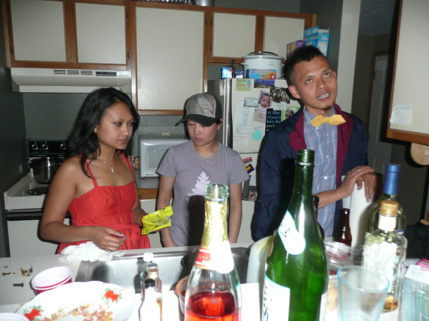 Zwei Frauen und ein Mann stehen in einer Küche mit Flaschen auf dem Spülenboden, einem Herd und einem Kühlschrank im Hintergrund.