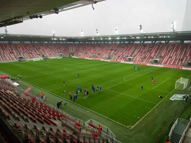 Ein Fußballfeld in einem Stadion mit Menschen, Stühlen, Geländern, Treppen und Lichtern unter einem offenen Himmel.