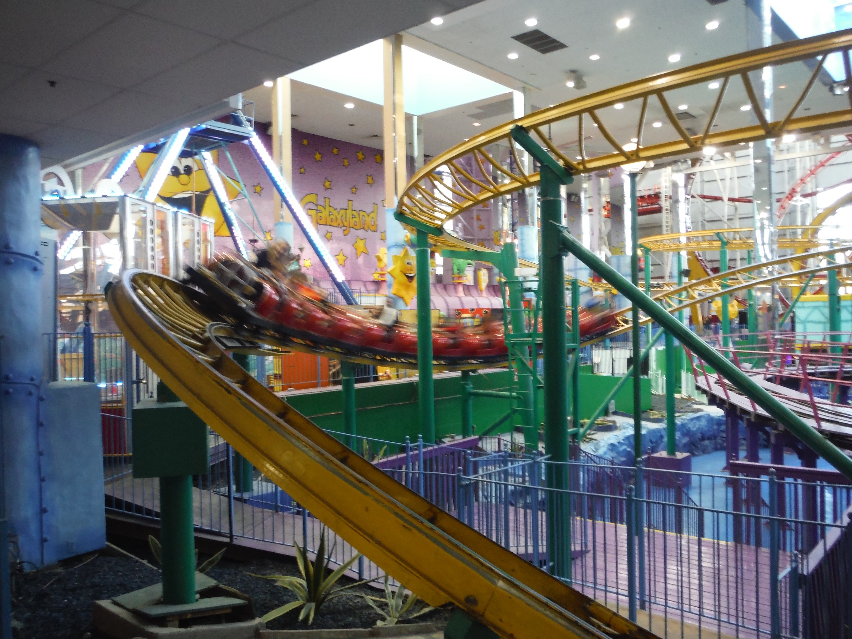 Großer Indoor-Freizeitpark mit Achterbahn, umgeben von Geländern, Säulen und Pflanzen, Deckenbeleuchtung und einigen Fahrgästen.