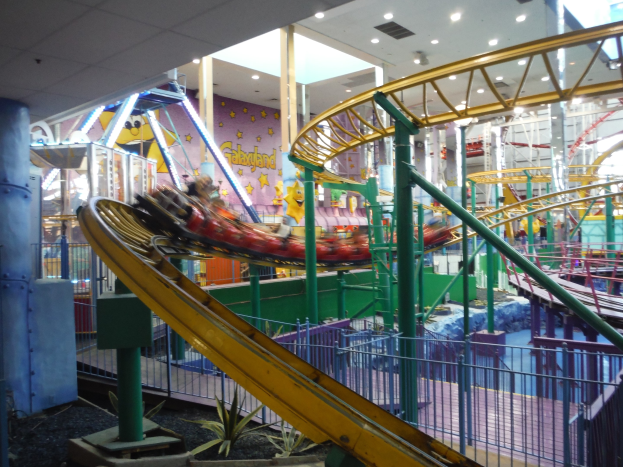 Großer Indoor-Freizeitpark mit Achterbahn, umgeben von Geländern, Säulen und Pflanzen, Deckenbeleuchtung und einigen Fahrgästen.
