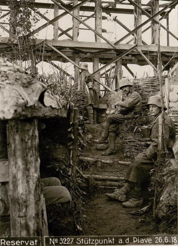 Eine Gruppe deutscher Soldaten in Helmen sitzt in einer Grube während des Zweiten Weltkriegs, umgeben von hölzernen Pfählen und Pflanzen, mit Text am unteren Rand des Schwarz-Weiß-Bildes.