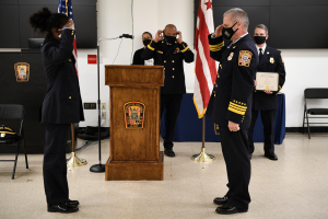 Eine Gruppe uniformierter Personen in Masken steht vor einem Podium mit Mikrofon, hält ein Zertifikat während einer Vereidigung eines neuen Feuerwehrchefs, umgeben von Fahnen, Stühlen, Tischen und einer Wand.