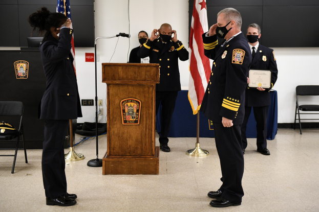 Eine Gruppe uniformierter Personen in Masken steht vor einem Podium mit Mikrofon, hält ein Zertifikat während einer Vereidigung eines neuen Feuerwehrchefs, umgeben von Fahnen, Stühlen, Tischen und einer Wand.