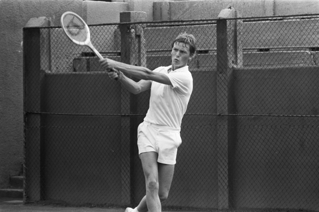 Mann in wei├čem Tennisoutfit schwingt einen Schl├Ąger auf einem Tennisplatz, umgeben von einem Zaun und einer Wand, in einem Schwarz-Wei├č-Bild.