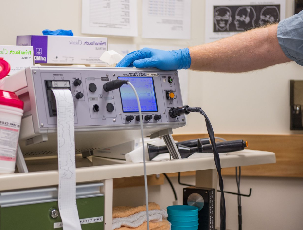 Eine Person in blauen Handschuhen arbeitet an einem medizinischen Gerät auf einem Tisch, umgeben von verschiedenen Gegenständen, mit Papieren an der Wand in einem Krankenhaus.