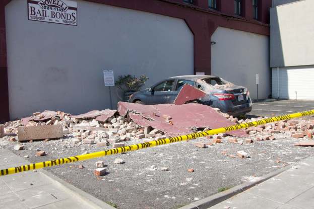 Eine gebrochene Wand mit verstreuten roten Ziegeln, ein geparktes Auto, eine Topfpflanze und ein Gebäude mit einer 'Kautionskaution'-Aufschrift auf einem Schild.
