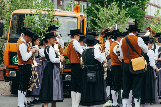 Gruppe von Menschen in traditioneller bayrischer Tracht, die eine Straße entlangmarschieren, Musikinstrumente spielen und Taschen tragen, mit Bäumen und Gebäuden im Hintergrund und einem geparkten Fahrzeug am Straßenrand.
